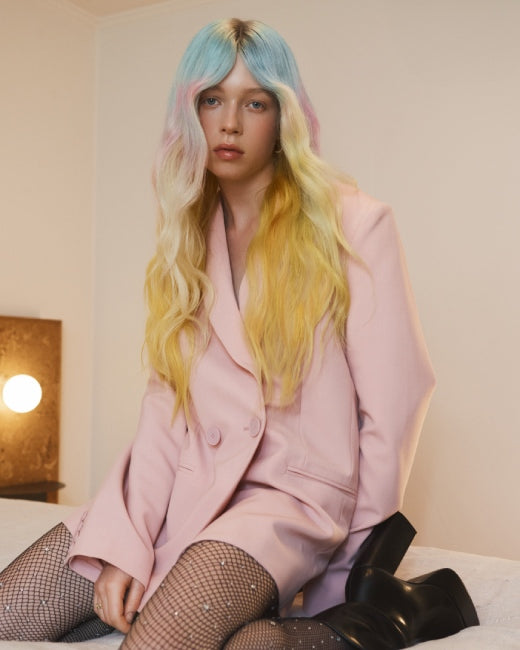 Person with pastel-colored hair wearing a pink suit sitting on a bed.
