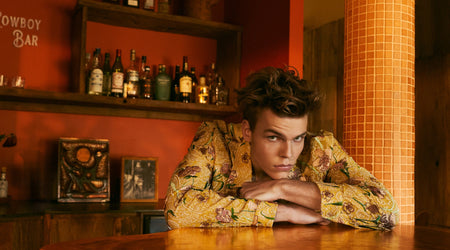 Man in a patterned shirt leaning on a bar counter with shelves of bottles in the background